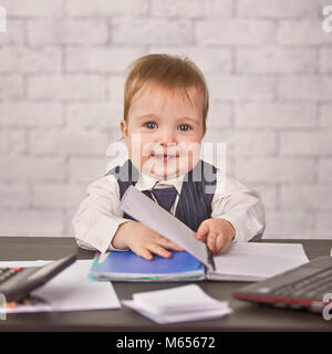 A cute blond child in a business suit is sitting at the desk. Infant broker with items for work Stock Photo