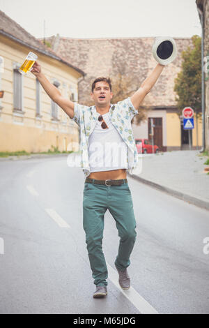 Portrait of cheerful drunk guy walking on the street and singing with hands up. He is holding stein full of beer and hat. Stock Photo