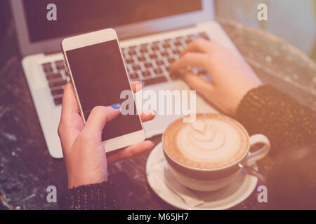 Businesswoman using blank screen smart phone and laptop on wooden table with cup of latte macchiato in cafe Stock Photo