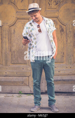 Cheerful hipster guy holding and looking at smart phone against old retro door background Stock Photo