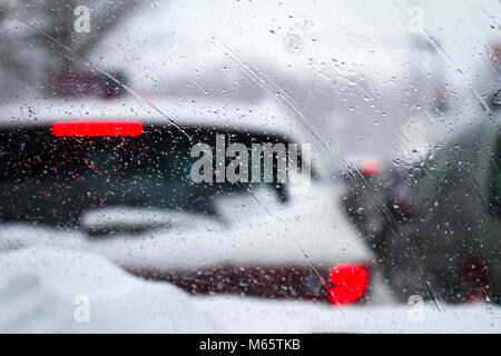 closeup of snowflakes and drizzle on windshield car driving through heavy winter city highway traffic with rear red lights of cars in front Stock Photo