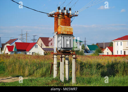 Old rusty electrical distribution transformer with cooling fins in the village. Against the backdrop of village houses. Stock Photo