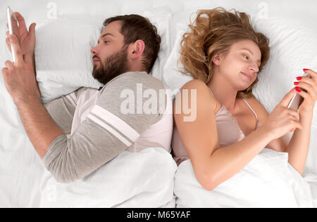 Young couple in bed using phone lying backs to each other. Stock Photo