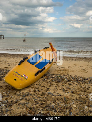 Southwold, England. A view from the beach looking out towards the sea.  A lifeguard's surfboard, from the RNLI, rests on the sand and stones ready to be launched out to sea at a moments notice.  A buoy can be seen floating in the sea close to the end of Southwold pier.  The sea is calm and it's a bright, but cloudy afternoon. Stock Photo