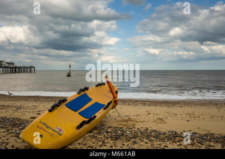 Southwold, England. A view from the beach looking out towards the sea.  A lifeguard's surfboard, from the RNLI, rests on the sand and stones ready to be launched out to sea at a moments notice.  A buoy can be seen floating in the sea close to the end of Southwold pier.  The sea is calm and it's a bright, but cloudy afternoon. Stock Photo