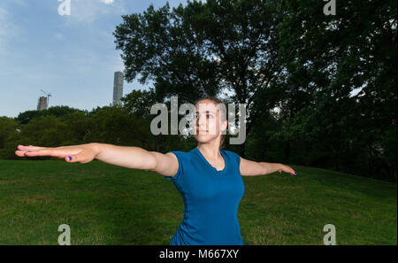 Young Caucasian Woman Practices Yoga In Central Park NYC While Wearing A Blue Top, Black Leggings and Barefoot Stock Photo