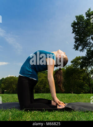 Young Caucasian Woman Practices Yoga In Central Park NYC While Wearing A Blue Top, Black Leggings and Barefoot Stock Photo