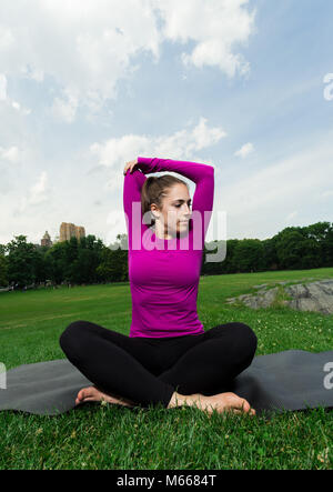 Young Caucasian Woman Practices Yoga In Central Park NYC While Wearing A Magenta Top, Black Leggings and Barefoot Stock Photo