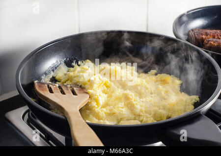 Scrambled eggs and vegetarian sausages, cooking in black frying pans on top of stove with wooden spatula and rising smoke. White tiled wall in backgro Stock Photo