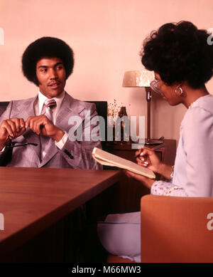 1970s AFRICAN AMERICAN STENOGRAPHER TAKING SHORTHAND DICTATION FROM AFRICAN AMERICAN BUSINESSMAN SEATED AT OFFICE DESK - ko697 HAR001 HARS COMMUNICATION CAREER TEAMWORK TWO PEOPLE INFORMATION TABLET FEMALES JOBS GROWNUP SEATED COPY SPACE HALF-LENGTH LADIES GROWN-UP INDOORS PROFESSION NOSTALGIA WORK PLACE 25-30 YEARS 30-35 YEARS WHITE COLLAR SKILL OCCUPATION SELLING SKILLS HIS STENOGRAPHER STYLES AFRICAN-AMERICANS AFRICAN-AMERICAN CAREERS BLACK ETHNICITY AFRICAN AMERICANS AFRICAN AMERICAN FASHIONS COOPERATION SALESMEN DICTATION MALES MID-ADULT MID-ADULT MAN MID-ADULT WOMAN SHORTHAND STENO PAD Stock Photo