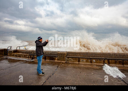 A man taking pictures of the crashing waves on the promenade wall at Colwyn Bay, Wales as a result of the Beast from the East and Storm Emma creating huge destructive waves along the coastline Stock Photo