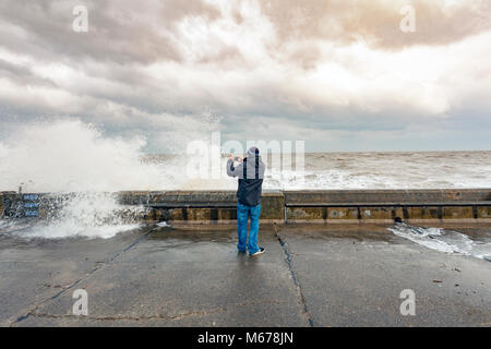 A man taking pictures of the crashing waves on the promenade wall at Colwyn Bay, Wales as a result of the Beast from the East and Storm Emma creating huge destructive waves along the coastline Stock Photo
