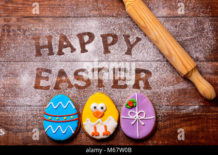 high-angle shot of a wooden table sprinkled with icing sugar or flour where you can read the text happy easter, some cookies decorated as ornamented e Stock Photo