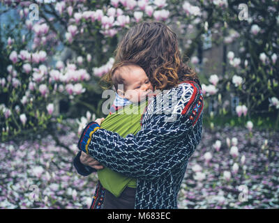 A young mother with her crying baby in a sling is standing by a magnolia tree Stock Photo