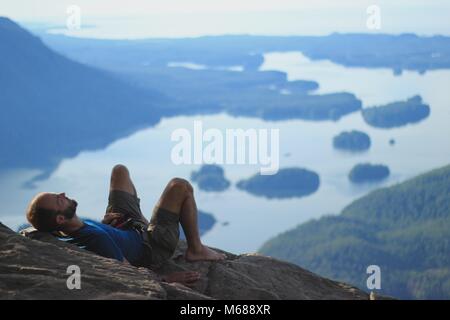 A man laying high above  on the edge of a mountain o a rock, lhaving a rest in front of the nature in front of him, green forest islands and ocean Stock Photo