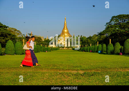 A young couple is walking on a meadow in front of the golden Shwedagon pagoda, the most sacred pagoda of Myanmar, located on a hill in the centre of t Stock Photo