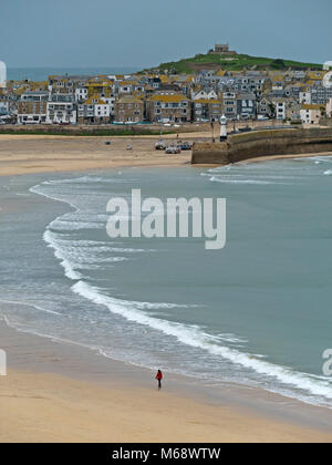 Lone figure walking on empty sandy beach by the sea with the harbour and seaside town of St. Ives in the distance, Cornwall, England, UK Stock Photo