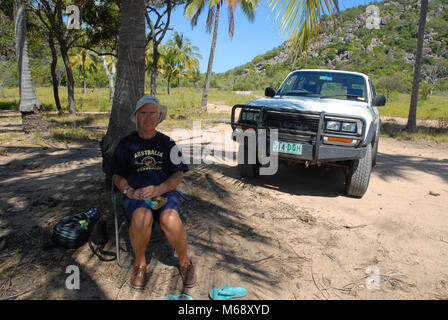 Senior man sitting under a coconut palm with 4WD off-road vehicle behind him, Radical Bay, Magnetic Island, Queensland, Australia Stock Photo
