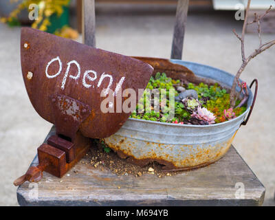 Rustic country Open sign, written in chalk on a rusty vice grip clamp plate. Sitting on an old sodden chair, and supported by a rusted out planter. Stock Photo