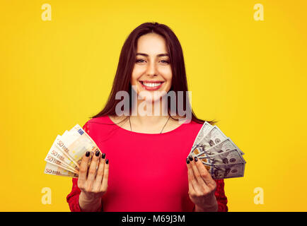 Young smiling woman excited with win and showing dollar and euro bills smiling at camera on yellow background Stock Photo