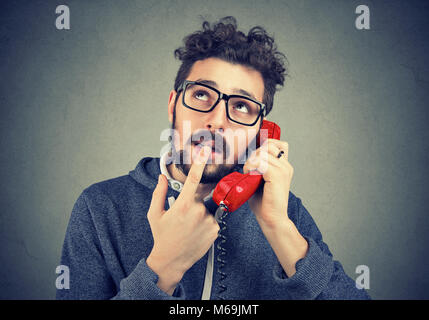 Young casual man looking up in confusion while talking on telephone against gray backdrop. Stock Photo