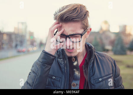 Young casual man frowning from headaches and noise while standing on urban background. Stock Photo