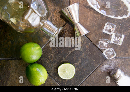 Ingredients for margarita cocktail with alcohol bottle, limes, salt and ice cubes with salt rimmed glass on granite. Stock Photo
