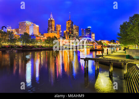 Blue sunrise in Melbourne city CBD on Yarra river banks looking at Walker foot bridge and high-rise towers above Flinders Station. Bright lights refle Stock Photo
