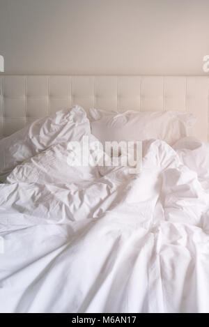 Unmade bed with plain white bed linen in a close up view of a rumpled duvet, pillows and a padded headboard Stock Photo