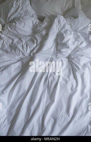 Unmade bed with plain white bed linen in a close up view of a rumpled duvet, pillows and a padded headboard Stock Photo