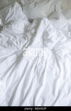 Unmade bed with plain white bed linen in a close up view of a rumpled duvet, pillows and a padded headboard Stock Photo