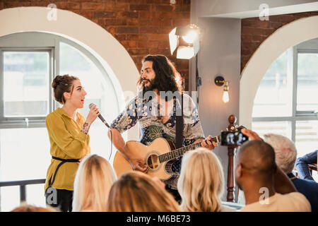 Wedding guests are enjoying some acoustic entertainment. There is a man playing guitar and a woman singing and everyone is watching and taking videos. Stock Photo