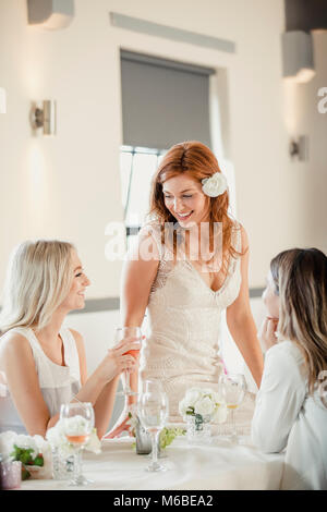Beautiful bride is socialising with some of her friends who are guests at her wedding. They are sat at a table, drinking wine as they talk. Stock Photo