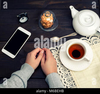 two female hands lie on a wooden table, next to a cup of tea and a smartphone with a black screen, top view Stock Photo