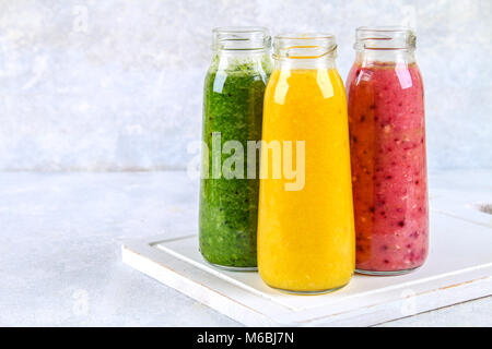Green, yellow, purple smoothies in bottles of berries, greens, oranges on a white board on a gray table Stock Photo