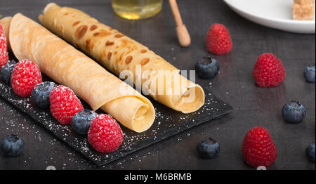 rolled pancakes with fresh strawberry’s and raspberry’s and blueberry’s on a black stone plate on a dark stone table with honey and white sugar and co Stock Photo