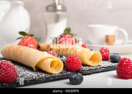 rolled pancakes with fresh strawberry’s and raspberry’s and blueberry’s on a black stone plate on a white wooden table with honey and white sugar and  Stock Photo
