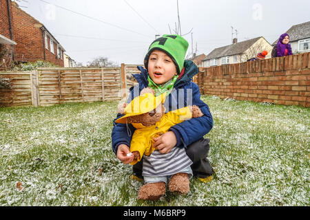 A young boy plays with his teddy bear in the front garden in the snow. Both the child and the toy are dressed in winter hats and coats. Stock Photo