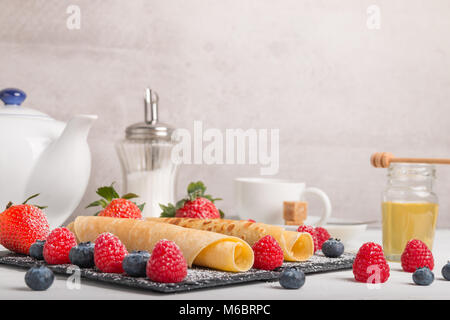rolled pancakes with fresh strawberry’s and raspberry’s and blueberry’s on a black stone plate on a white wooden table with honey and white sugar and  Stock Photo