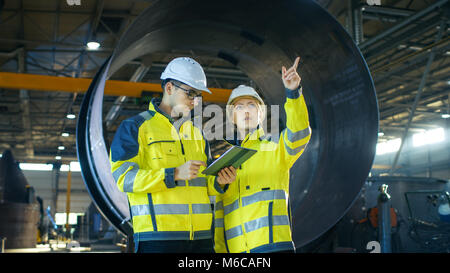 Male and Female Industrial Engineers Have Discussion while Using Tablet Computer. They Work in a Heavy Industry Manufacturing Factory. Stock Photo