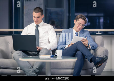 Two young businessmen sitting in office lobby and using laptop and digital tablet device Stock Photo
