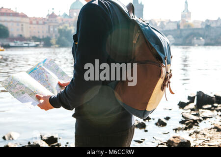 A young tourist man with a backpack standing next to the river Vltava in Prague looks at the map and admires the architecture of the city. Charles Bridge is near. Travel around the Czech Republic. Stock Photo