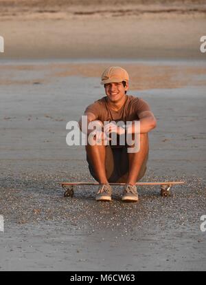 Hip, young man taking a break, sitting on skateboard on beach to watch sunset, making "shaka" sign (chill out), in San Diego, California, USA Stock Photo