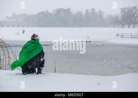 Celbridge, Kildare, Ireland. 02 Mar 2018: Photographer in green cape watching and photographing mallard ducks standing on a frozen pond at the covered in snow Castletown Park in Celbridge. Beast from the east followed by Storm Emma brought snow and high winds paralyzing the country   and forcing Met Éireann to issue the red warning alert across Ireland. Stock Photo