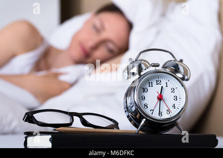 Close-up Of Eyeglasses And Alarm Clock On Table In Front Of Sleeping Woman Stock Photo