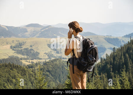 Girl with tourist rucksack on a long hiking walk talks on smartphone up in the hills Stock Photo