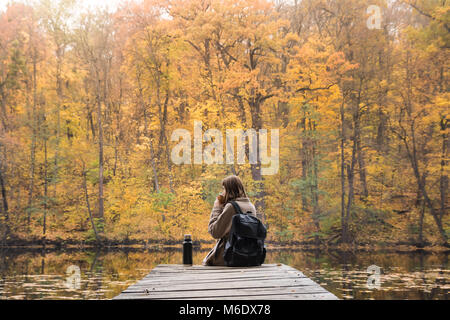 Young female person hiking at nature park rests at riverbank on gold autumn day and enjoys beautiful october scenery having a hot drink from thermos Stock Photo