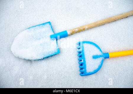 A child's toy spade and rake are covered in snow having been left outside on a garden table before it snowed. Stock Photo