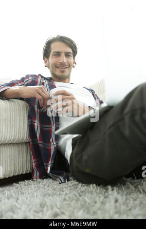 young man with laptop holding a cup sitting on the floor near the sofa Stock Photo