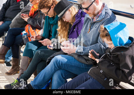 Family sitting on a bench, all of whom are simultaneously looking at their mobile phone / device / devices, texting or surfing / using web / internet. Stock Photo